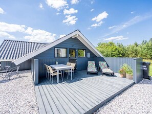 A modern house with a sloped roof, a wooden deck, a dining area with chairs and a table, and two sun loungers.