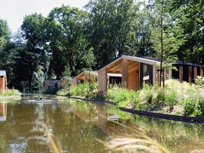 A row of wooden cabins with a pond in the foreground, surrounded by lush greenery. - Forest Chalet Escape (Maarn)