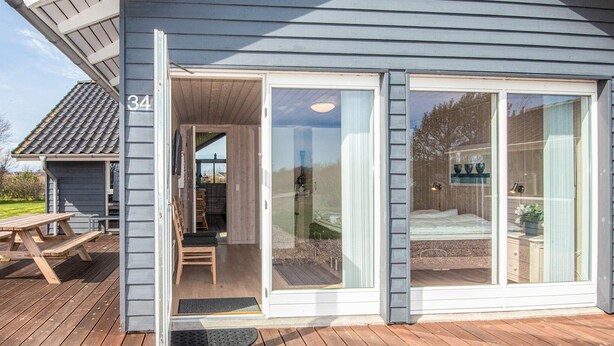 A house with a number 34, a wooden deck, a sliding glass door, and a visible interior with a bed and a window. - Holiday Home in Tarm (Tarm)
