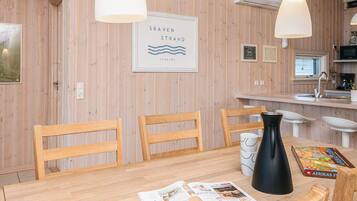 A wooden interior with a dining table, chairs, a black vase, a magazine, and a framed sign reading 'SKAVEN STRAND DANMARK'.