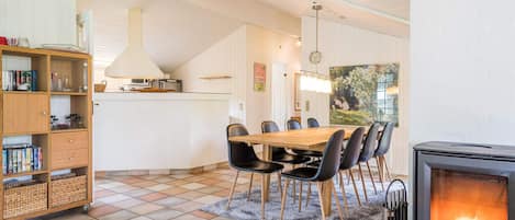 A dining area with a wooden table and chairs, a kitchen in the background, and a fireplace with a visible fire.