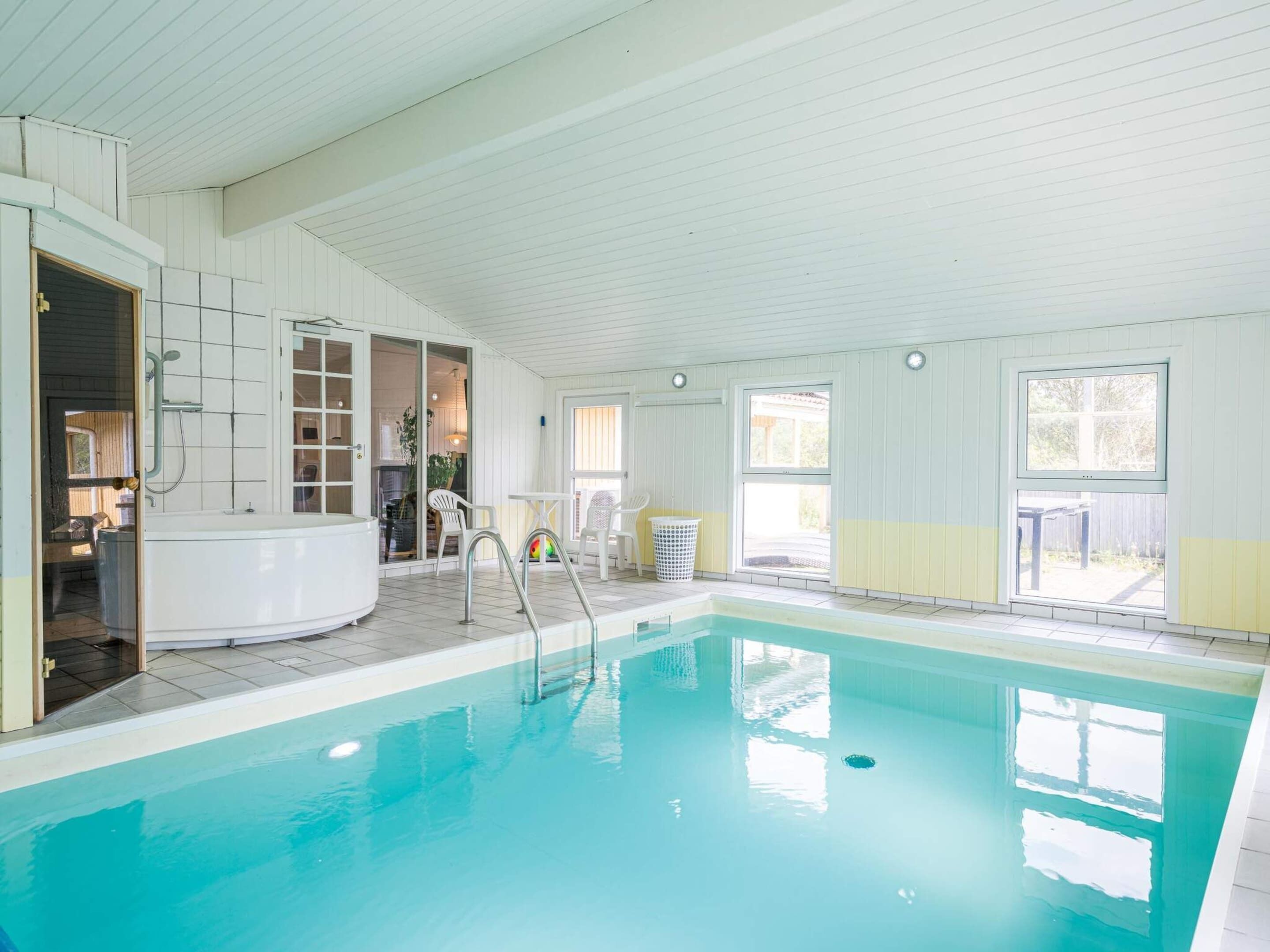 En indendørs swimmingpool med jacuzzi, et lyst rum med bord og stole, og udsigt til det fri.