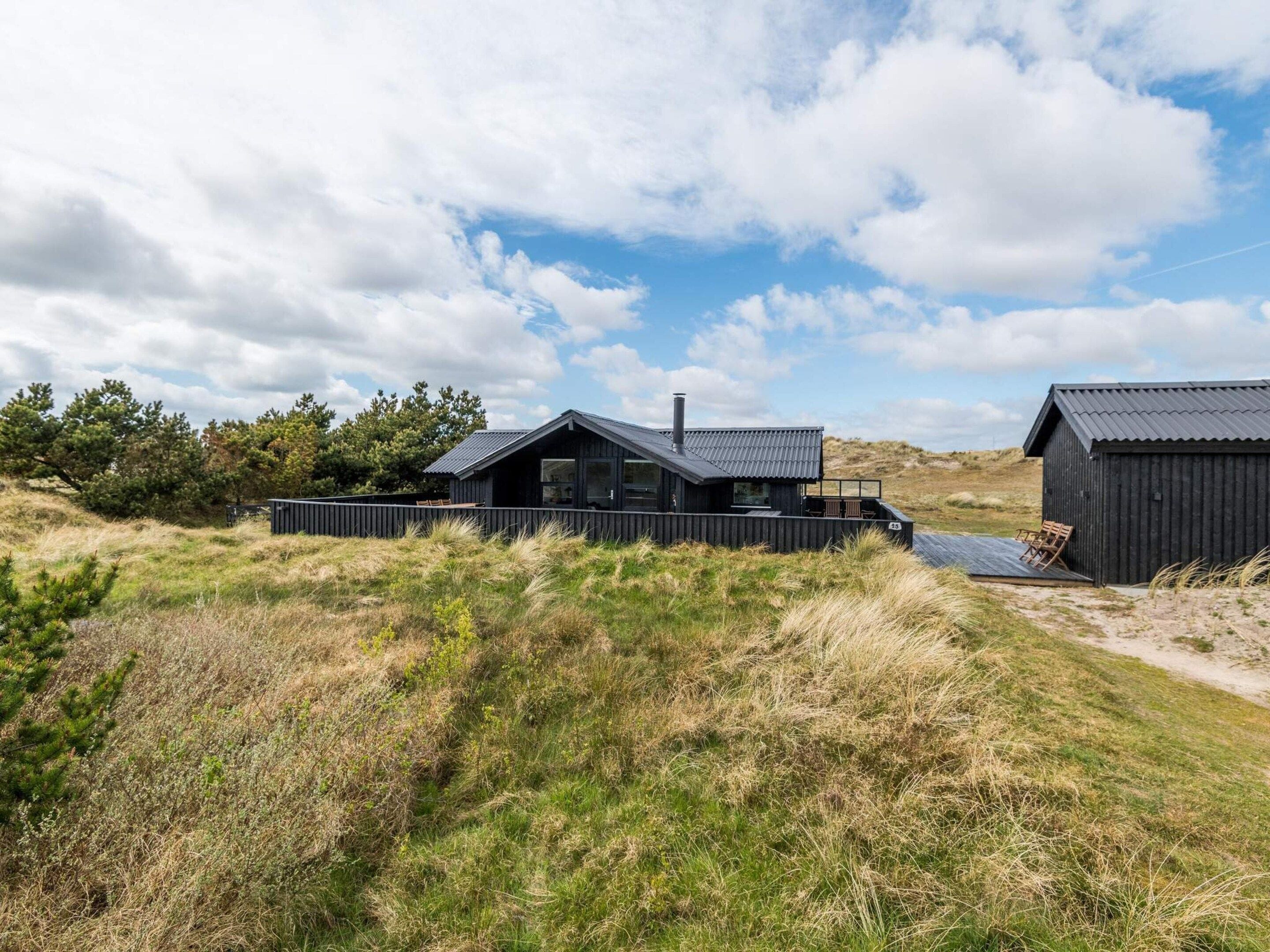 Une cabane moderne, de couleur noire, avec un toit en pente, une terrasse et un petit espace extérieur aménagé pour s’asseoir.