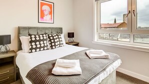 Ein Hotelzimmer mit Bett, Fenster mit Stadtblick, einem gerahmten Kunstwerk an der Wand und einem Nachttisch mit Lampe.