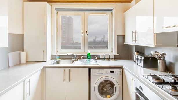Eine moderne Küche mit weißen Schränken, einer integrierten Waschmaschine, einem Fenster mit Blick auf die Stadt und einem Herd.