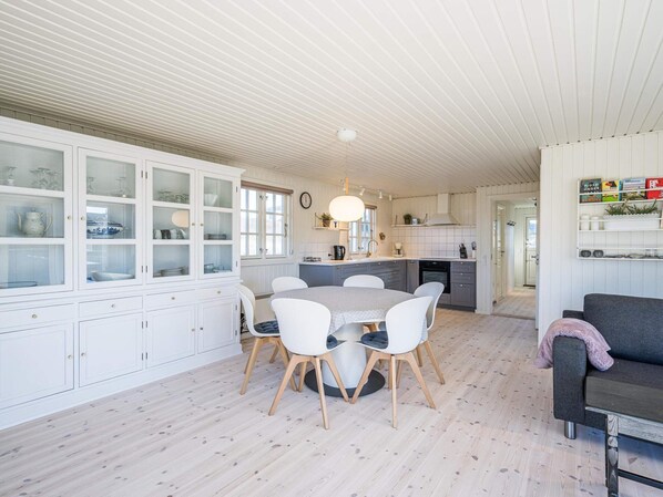 Eine moderne Küche mit weißen Schränken, ein Essbereich mit rundem Tisch und weißen Stühlen sowie ein graues Sofa in einem hellen, offenen Raum mit Holzboden.
