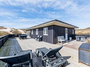 A wooden deck with outdoor furniture, a building with large windows, and a view of the dunes in the background.