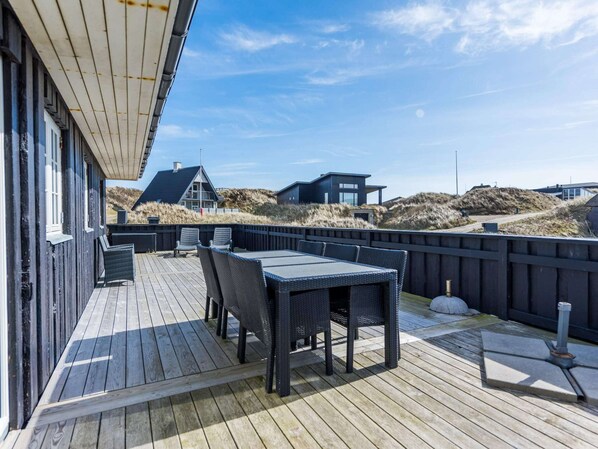 A wooden deck with outdoor furniture, a view of dunes, and a modern building in the background. - Seaside Gem in Sondervig-by Traum (Ringkobing)