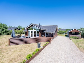 A modern cabin with a deck, a covered outdoor seating area, and a gravel driveway.