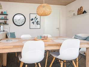 A dining area with a wooden table, white chairs, a woven pendant light, a wall clock, and a framed artwork.