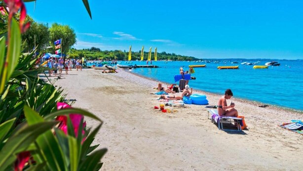 A beach with people sunbathing and swimming, boats in the water, and flags on the shore. - Holiday Apartment near Lake, Shared Swimming Pool (Lazise)