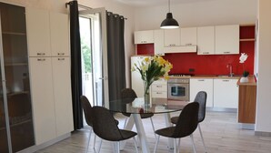 A modern kitchen with white cabinets, a red accent wall, and a dining area with a glass table and wicker chairs. - Holiday Apartment near Lake, Shared Swimming Pool (Lazise)