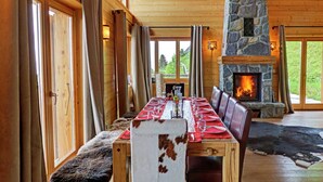 Un espace repas avec une longue table en bois dressée pour le repas, une cheminée en pierre et de grandes fenêtres offrant une vue sur l’extérieur.