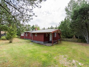Une petite cabane à un seul étage, dotée d’un porche, entourée d’une cour verdoyante et d’arbres.