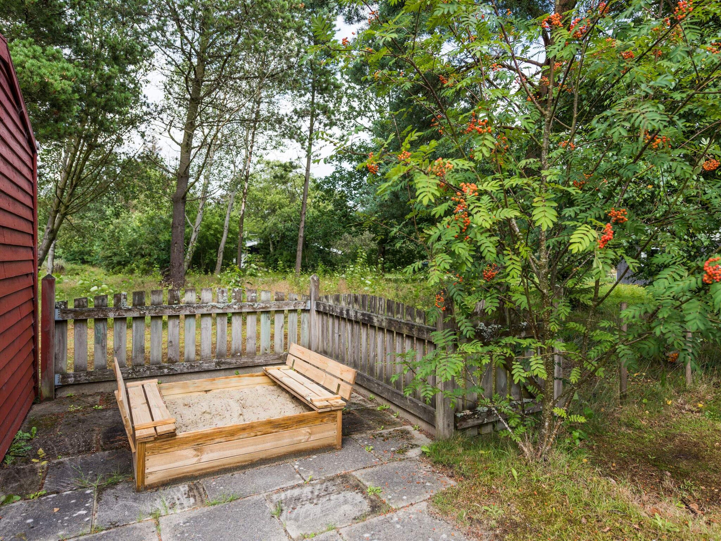 Un área de estar exterior de madera con un banco y una mesa, rodeada por una valla de madera y árboles.