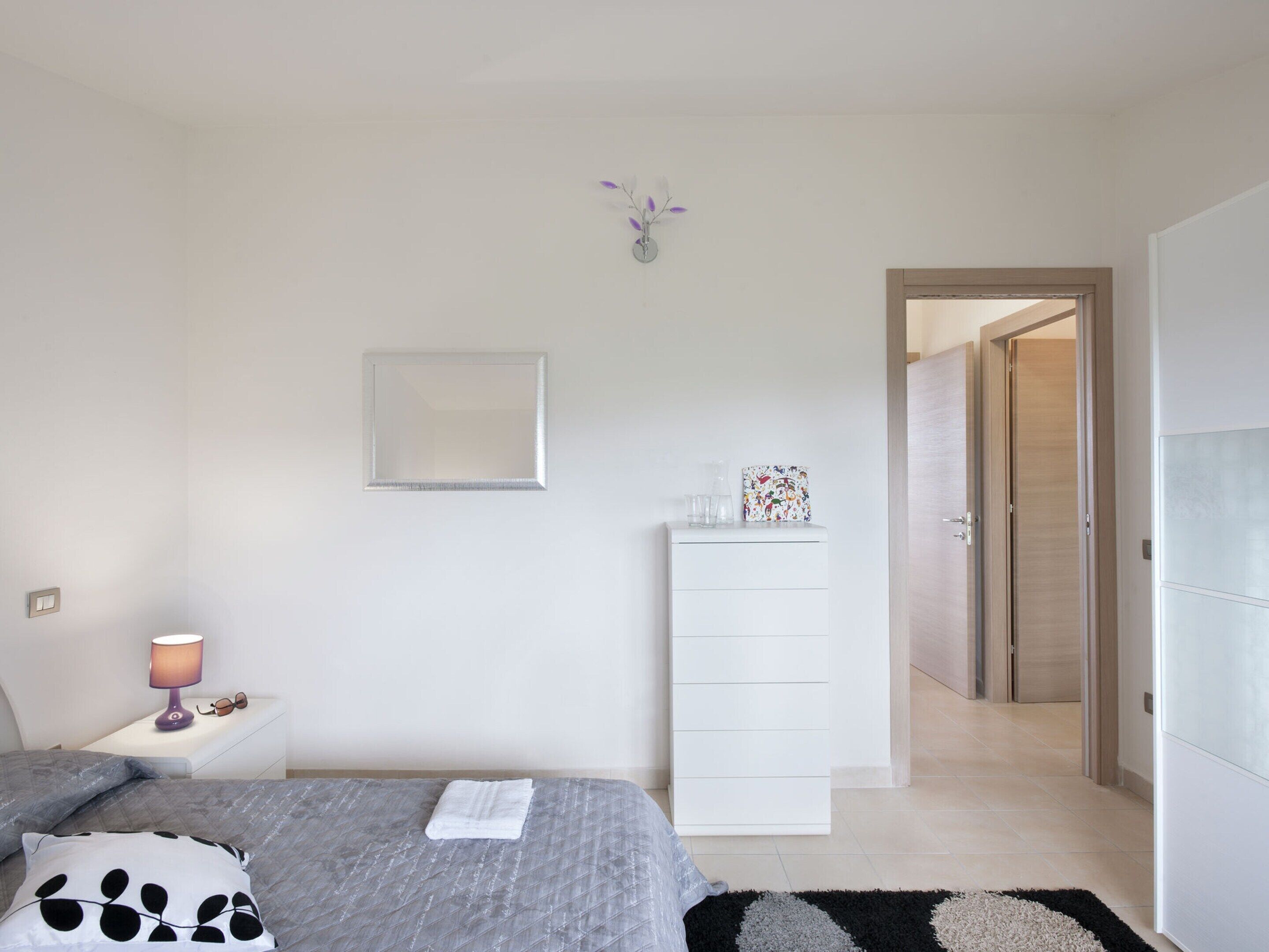 A bedroom with a bed, a nightstand with a lamp, a dresser, and a door leading to another room.