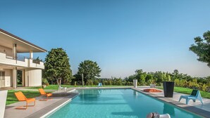 Una piscina moderna al aire libre con sillas de descanso y una mesa, rodeada de vegetación y un edificio con balcones.