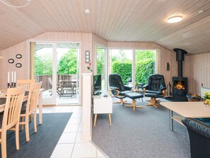 A cozy room with a wooden ceiling, a fireplace, a dining area with chairs, and a large window offering a view of greenery.
