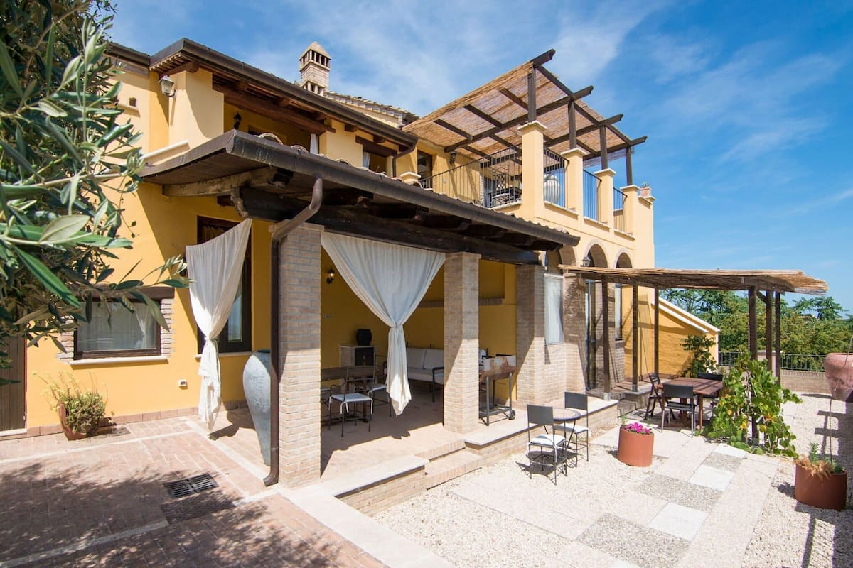 Ein gelbes Gebäude mit überdachter Terrasse, Außenbestuhlung und einer Pergola.