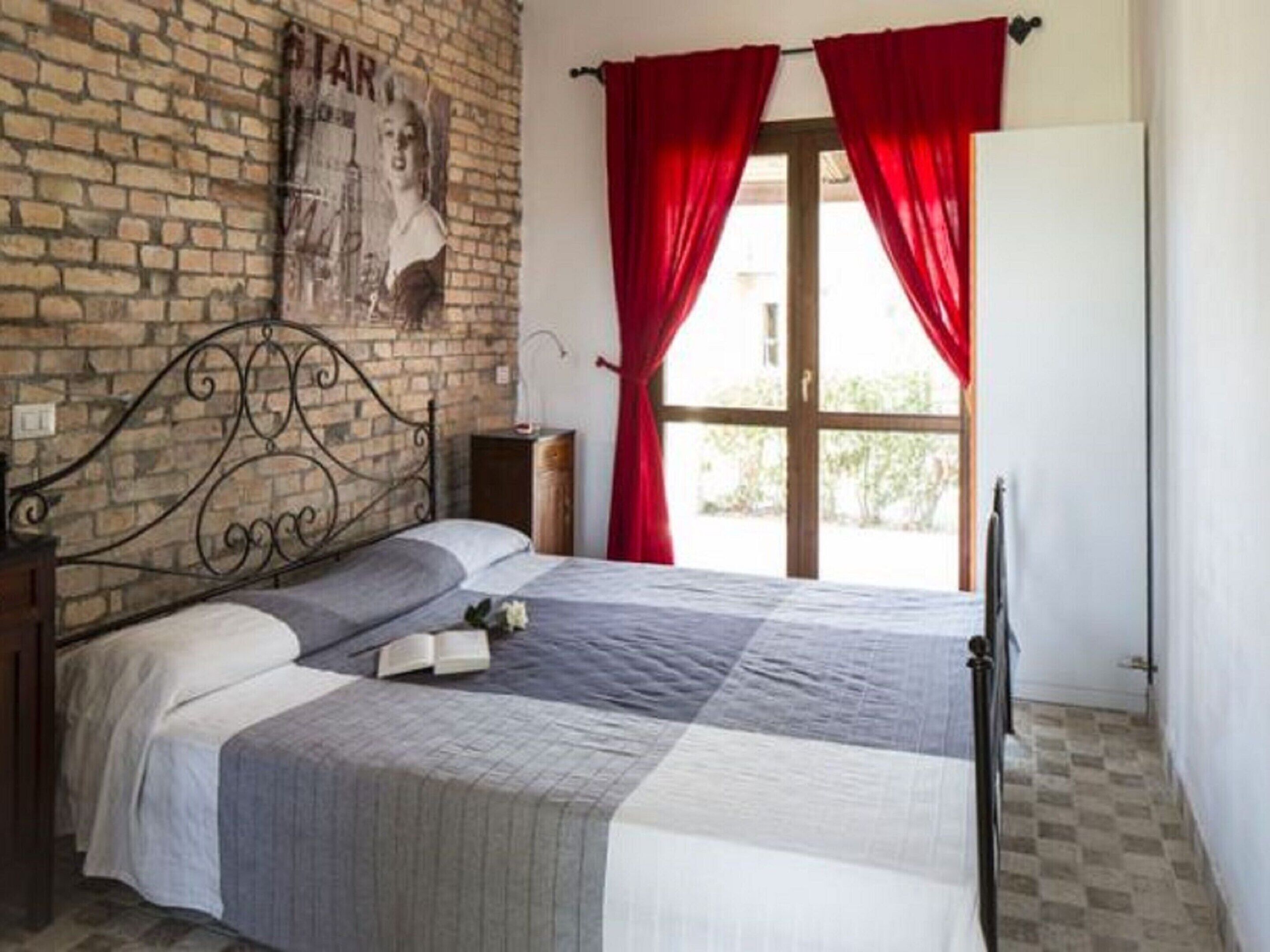 A bedroom with a brick wall, a metal bed frame, a checkered bedspread, and red curtains.