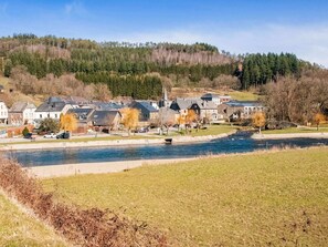 A riverside residential area with houses, a stone bridge, and a forested hill in the background.