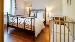 A bedroom with a metal bed frame, a white crib, a window with curtains, and a wall-mounted picture. - Timeless Cottage in Vresse-sur-semois With Terrace (Vresse-sur-Semois)