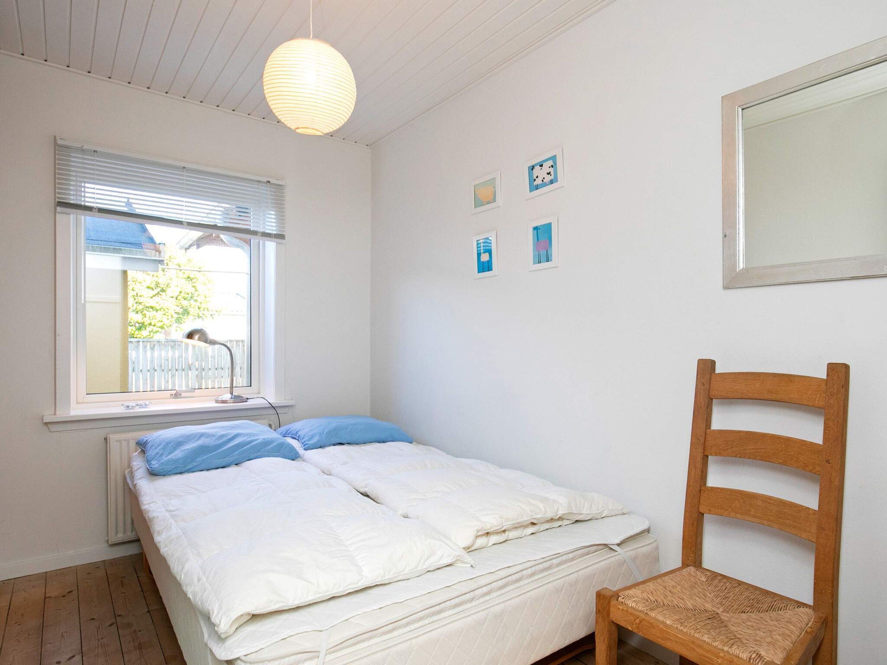Une chambre à coucher avec un lit, une chaise en bois, une fenêtre avec des stores et des œuvres d’art accrochées au mur.