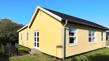 Una casa amarilla con techo negro, ventanas blancas y un pequeño porche con sillas.