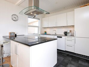 A modern kitchen with white cabinets, a black countertop, a stainless steel range hood, a clock, and a window providing natural light. - 8 Person Holiday Home in Skagen (Skagen)