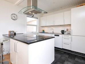 A modern kitchen with white cabinets, a black countertop, a stainless steel range hood, a clock, and a window providing natural light. - 8 Person Holiday Home in Skagen (Skagen)