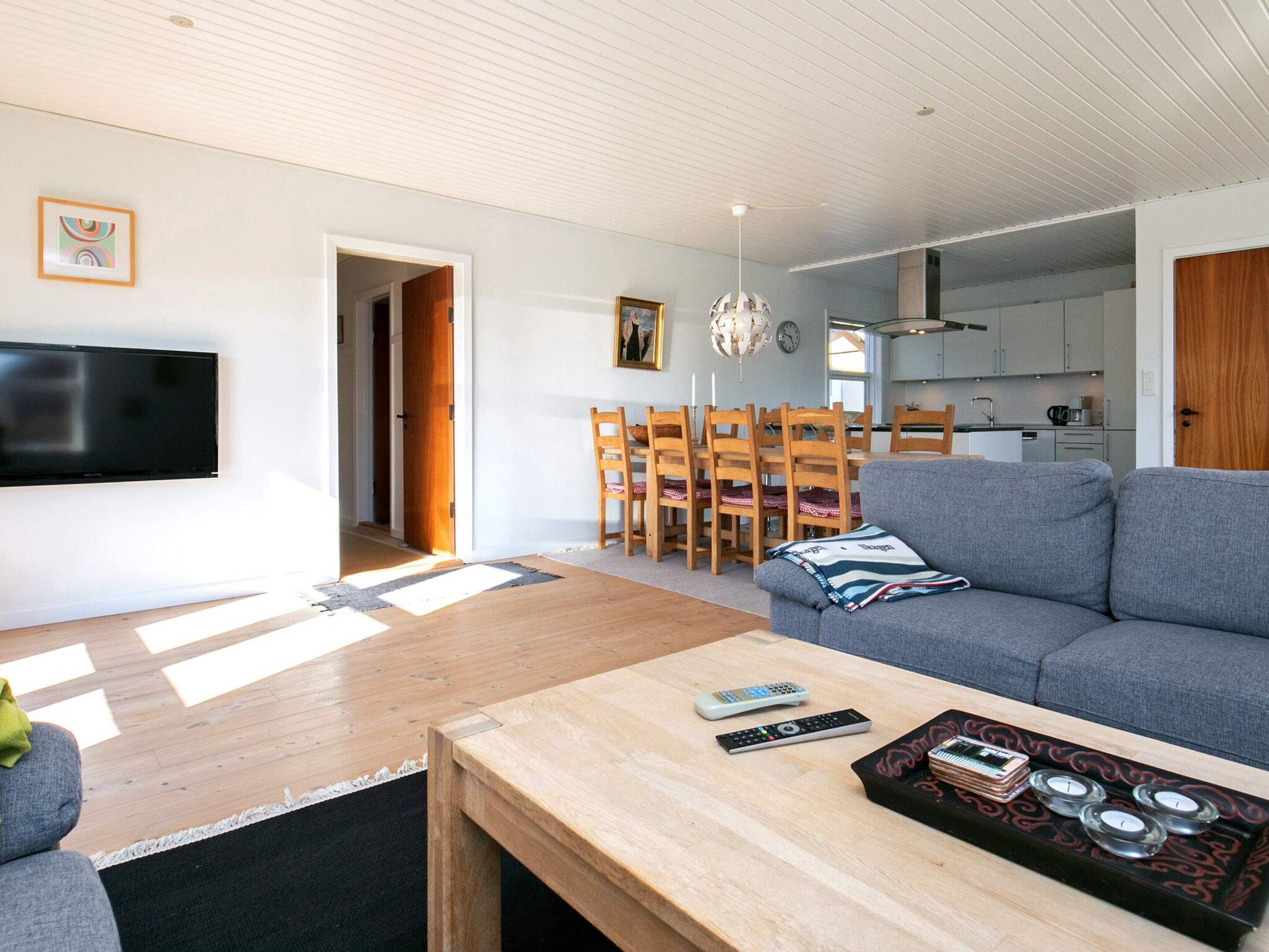 A modern living room with a grey sofa, wooden dining table, and a flat-screen TV mounted on the wall.