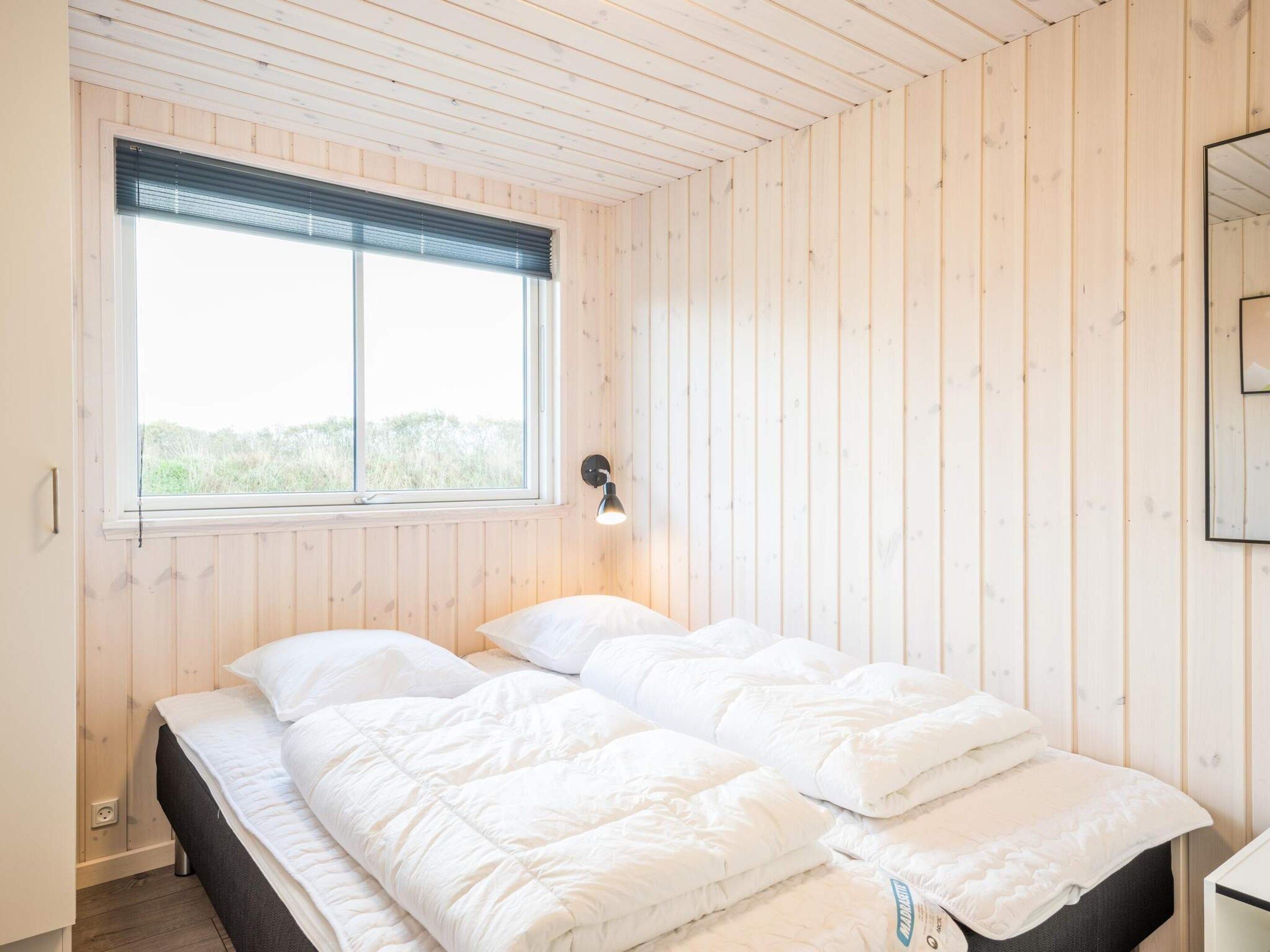 Ein Zimmer mit Holzwänden, einem Bett mit weißer Bettwäsche, einem Fenster mit Jalousien und einer Wandleuchte.