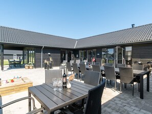 An outdoor patio area with multiple tables and chairs, a wooden table with a bottle and glasses, and a grill in the background. - 5 Star Holiday Home in Norre Nebel (Norre Nebel)