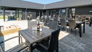 An outdoor patio area with multiple tables and chairs, a wooden table with a bottle and glasses, and a grill in the background. - 5 Star Holiday Home in Norre Nebel (Norre Nebel)