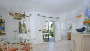 A spacious dining area with a glass table and white chairs, a white cabinet with a bird sculpture, and a view of the outdoors through a glass door. - Beautiful Villa Located Less Than Five Minutes Walk From the Old Village (Loulé)