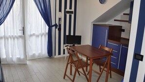 Un petit appartement moderne doté d’un coin repas avec une table et des chaises en bois, d’une kitchenette avec des placards bleus et d’une grande fenêtre ornée de rideaux bleus.