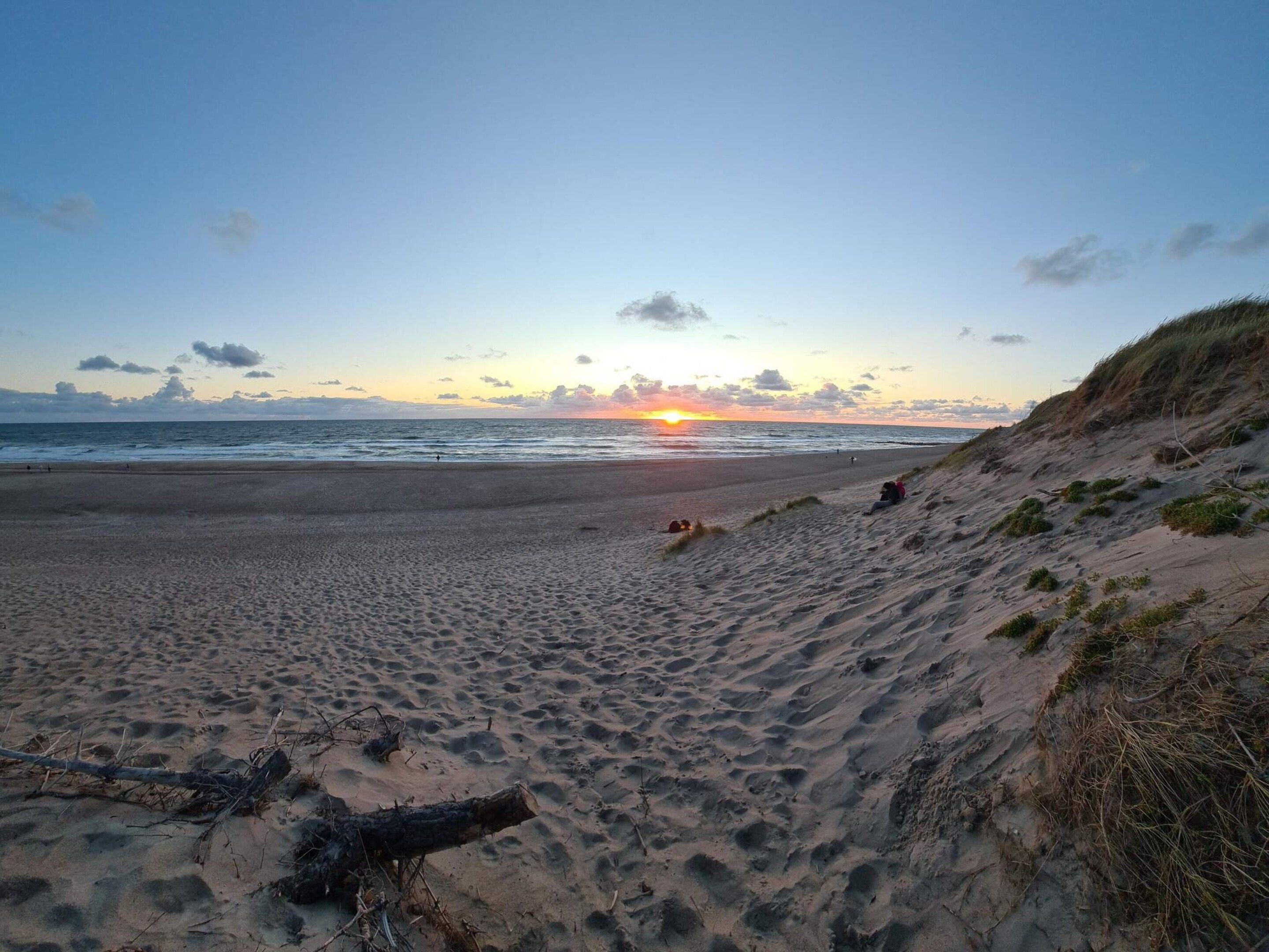A beach with a sunset, sandy terrain, and a grassy dune.