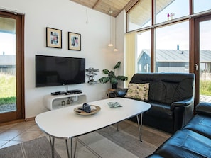 Una sala de estar moderna con un televisor de pantalla plana, un sofá de cuero negro, una mesa de centro blanca y un ventanal con vistas al exterior.