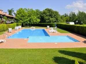 A rectangular outdoor swimming pool with two separate sections, surrounded by a brick patio. There are lounge chairs and a small table with a chair, and a hedge in the background.