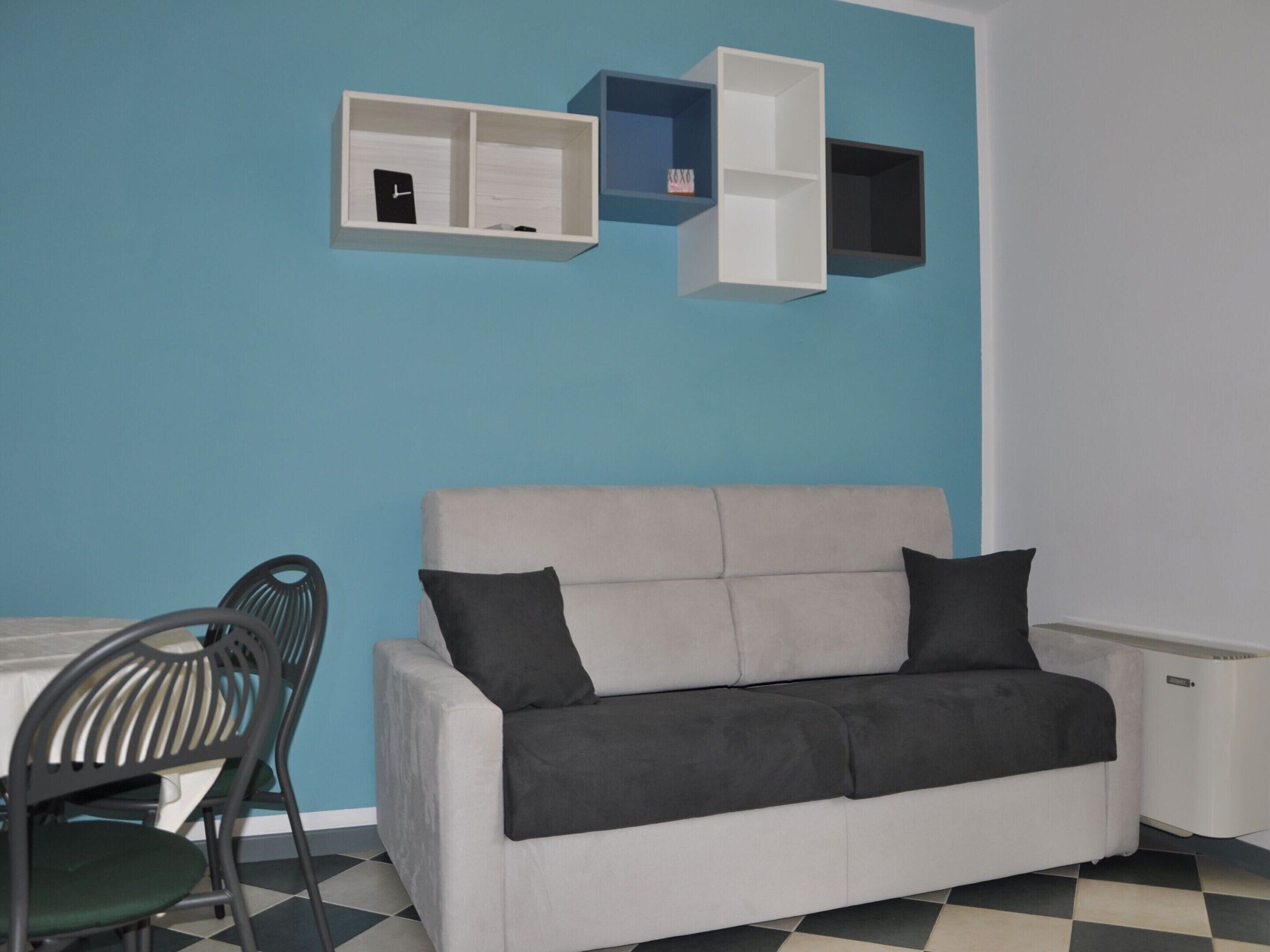 Una habitación con un sofá gris, dos cojines negros, una mesa blanca y una silla. Hay tres estantes blancos en la pared, uno con un reloj y otro con un objeto pequeño. El suelo tiene un estampado de cuadros.