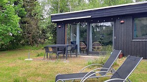 Un área de estar moderna al aire libre con sillones reclinables y una mesa, rodeada por un edificio de madera con ventanas amplias.