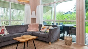 Ein modernes Wohnzimmer mit einem grauen Ecksofa, einem Holztisch und Blick ins Freie durch große Fenster.
