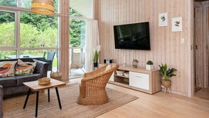 Ein modernes Wohnzimmer mit großem Fernseher, Korbmöbeln und Blick ins Grüne.