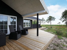 Una casa moderna con terraza de madera, mobiliario exterior negro y vistas del paisaje circundante.
