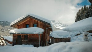 Une cabane en bois dont le toit est recouvert de neige, entourée d’un paysage enneigé et d’arbres, avec des montagnes en arrière-plan.