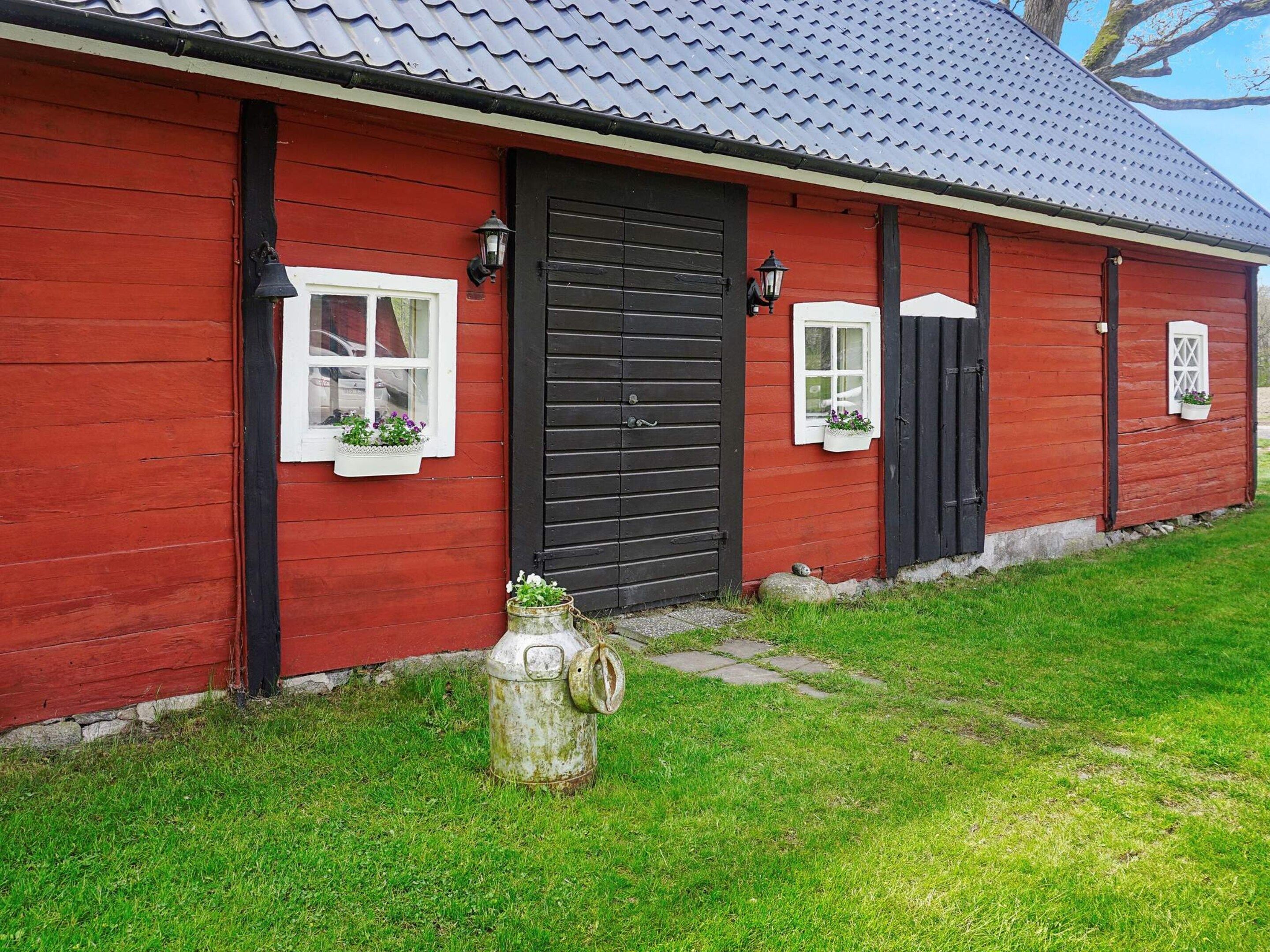 A red wooden building with a black door, two windows with white frames, and a small stone structure in front.
