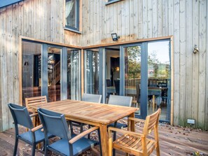 Una terraza de madera con una mesa grande y sillas, rodeada de puertas de cristal que dan a un espacio interior.