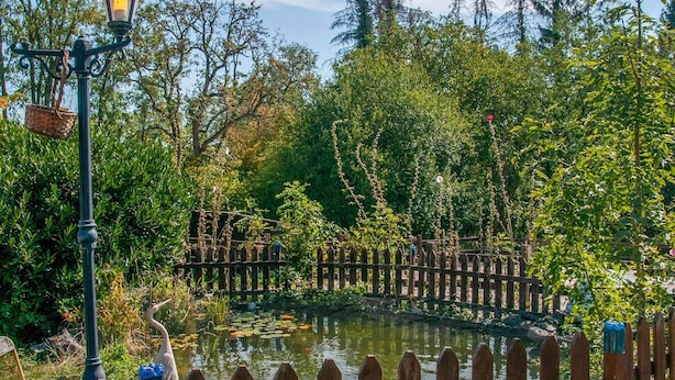 Une clôture en bois entoure un étang sur lequel se trouve un pont en bois, le tout étant bordé d’une végétation luxuriante et d’arbres.
