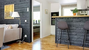 A modern apartment with a dark wood wall, a wooden floor, a kitchen with a microwave, and a seating area with a sofa and two bar stools. - 4 Star Holiday Home in Loftahammar (Loftahammar)