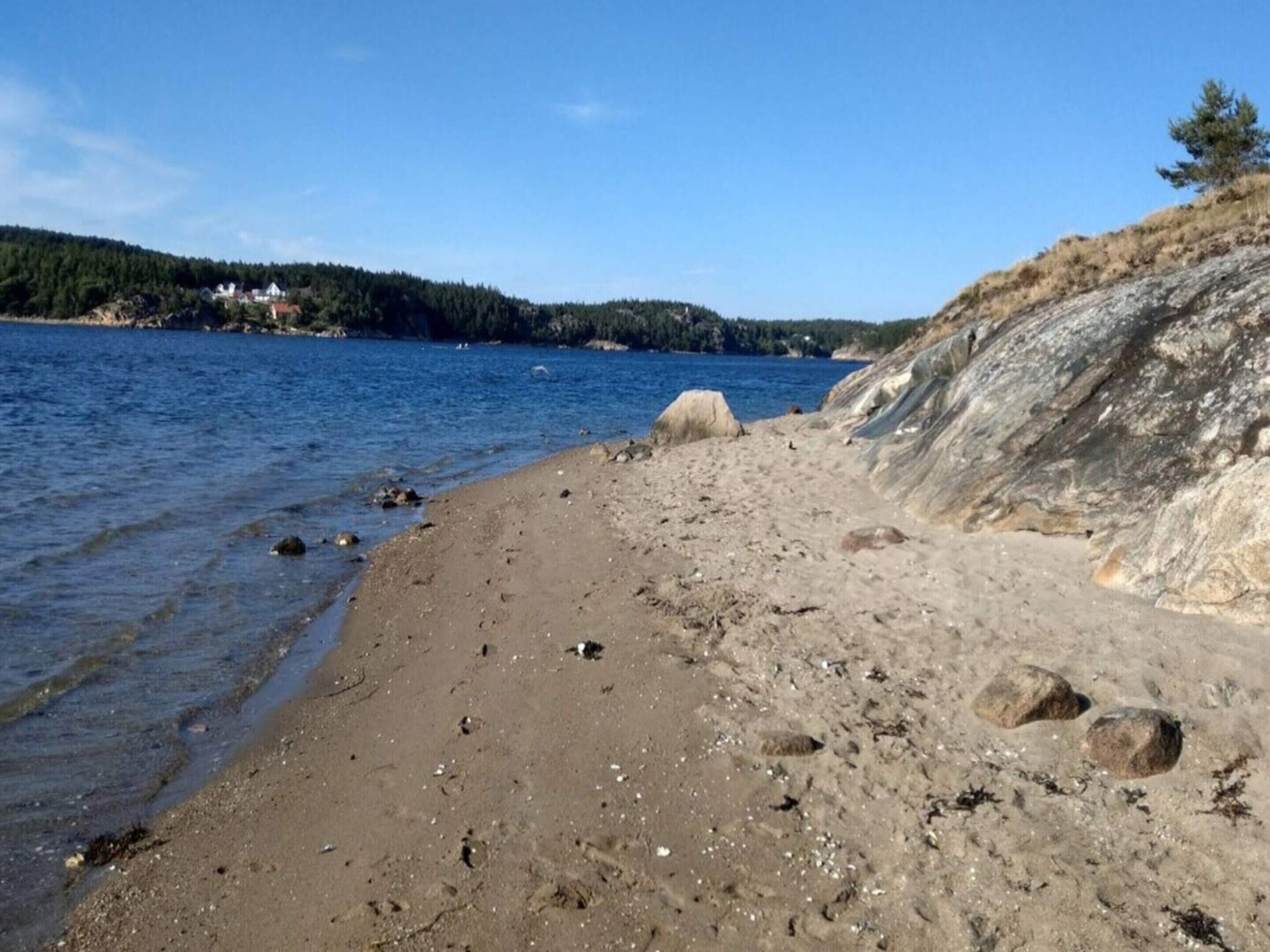 A sandy beach with clear water, a rocky shoreline, and a forested hill in the background.